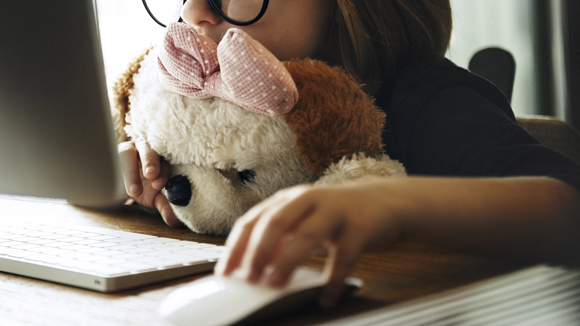Menina de óculos abraçando um urso de peluche enquanto utiliza um computador.